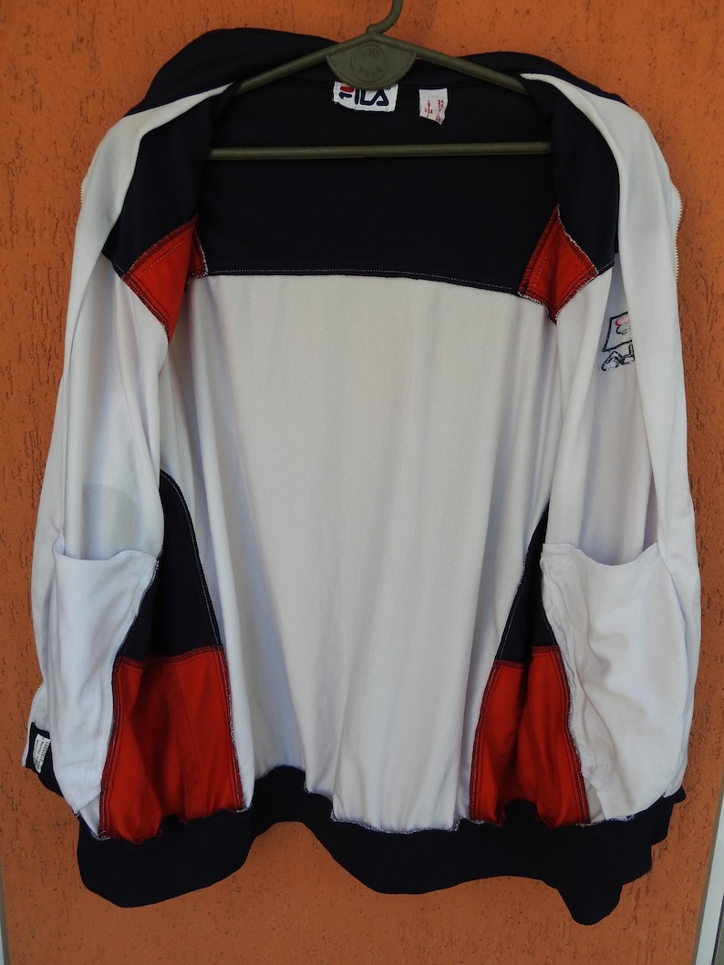 Puede incluir: Una chaqueta deportiva con cremallera Fila que presenta un dise&ntilde;o de bloques de color en blanco, azul marino y rojo. La chaqueta tiene un cuerpo principal blanco, hombros y dobladillo azul marino, y detalles rojos en los laterales.