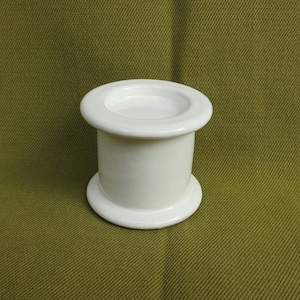 May include: A white ceramic candle holder with a cylindrical body and a wide, flat base and top. The candle holder is smooth and glossy, and it sits on a green surface. The design is simple and elegant.