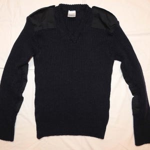 Puede incluir: Un suéter de cuello en V azul marino oscuro con textura de punto acanalado, mangas largas y parches de tela negra sólida reforzados en los hombros y codos. La prenda incluye una etiqueta blanca en el cuello con texto visible.