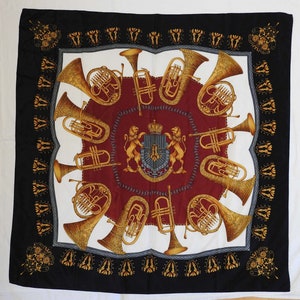 Könnte beinhalten: Quadratisches Tuch mit schwarzem Rand und einem zentralen Design mit Blechblasinstrumenten, einem Wappen und einem bordeauxroten Hintergrund. Die Instrumente sind goldfarben, und das Wappen zeigt eine Krone und einen Löwen.