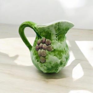 May include: A ceramic pitcher featuring a textured green leaf design with a raised cluster of purple grapes on the side. The pitcher has a curved handle and a wide spout, with a glossy finish that highlights the detailed botanical relief pattern.