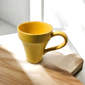 May include: A yellow ceramic mug with a curved handle. The mug has a slightly textured surface and a wide opening. The interior of the mug is a bright yellow, contrasting with the exterior. The mug sits on a wooden surface.