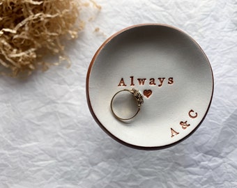 May include: White ceramic ring dish with a brown rim. The dish has the words "Always" and "A & C" inscribed on it. A gold ring with a heart-shaped stone is sitting in the dish.