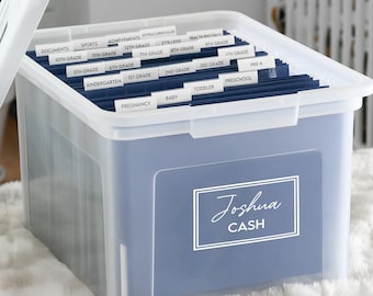 Op de afbeelding: Een doorschijnende plastic opbergdoos met een wit deksel, gevuld met donkerblauwe hangmappen met witte labels. De voorzijde van de doos is voorzien van een rechthoekig label met de witte tekst Joshua CASH binnen een dubbel omlijnd kader.
