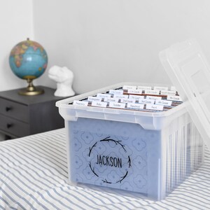以下が含まれることがあります： 青い模様のインサートと、リースデザインの「JACKSON」という名前が入った透明なプラスチック製の収納ボックス。ボックスにはラベル付きのファイルフォルダーが入っています。蓋は部分的に開いています。地球儀と小さなサイドテーブルが背景にあります。