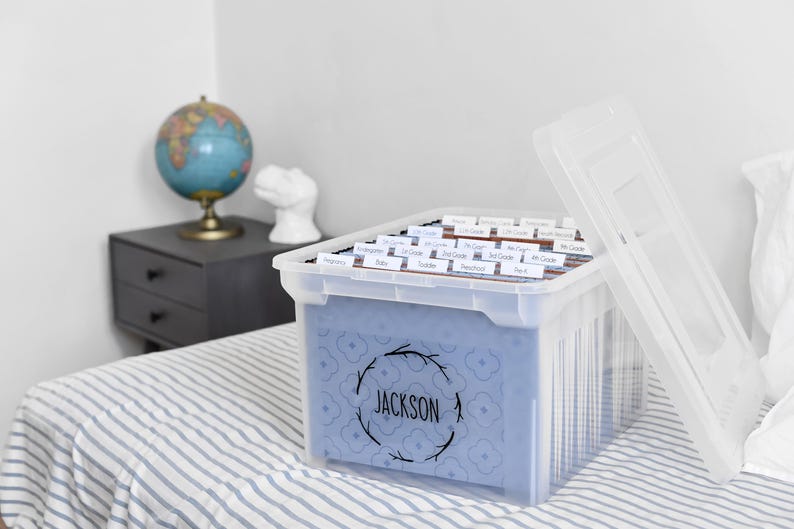 以下が含まれることがあります： 青い模様のインサートと、リースデザインの「JACKSON」という名前が入った透明なプラスチック製の収納ボックス。ボックスにはラベル付きのファイルフォルダーが入っています。蓋は部分的に開いています。地球儀と小さなサイドテーブルが背景にあります。