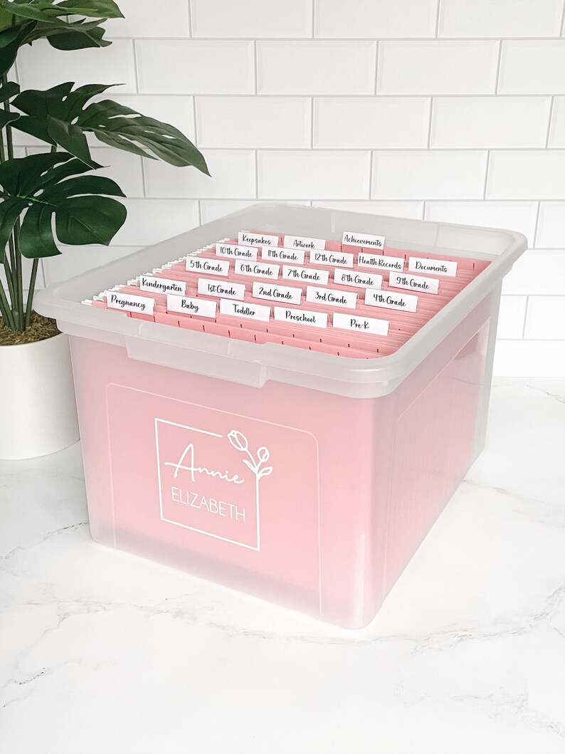 以下が含まれることがあります： 透明な蓋が付いたピンク色の収納ボックス。ラベル付きのファイルフォルダーでいっぱい。ラベルには「幼稚園」、「1年生」、「実績」などのカテゴリーが含まれています。ボックスの前面には「Annie Elizabeth」という名前が印刷されています。