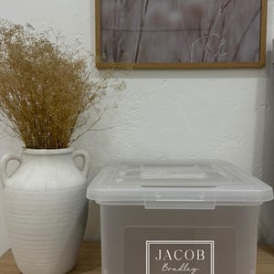 May include: A clear plastic storage bin with a gray front panel and lid. The front panel features the name "JACOB Bradley" in a white, elegant font within a white border. A white ceramic vase with handles holds dried brown plants.