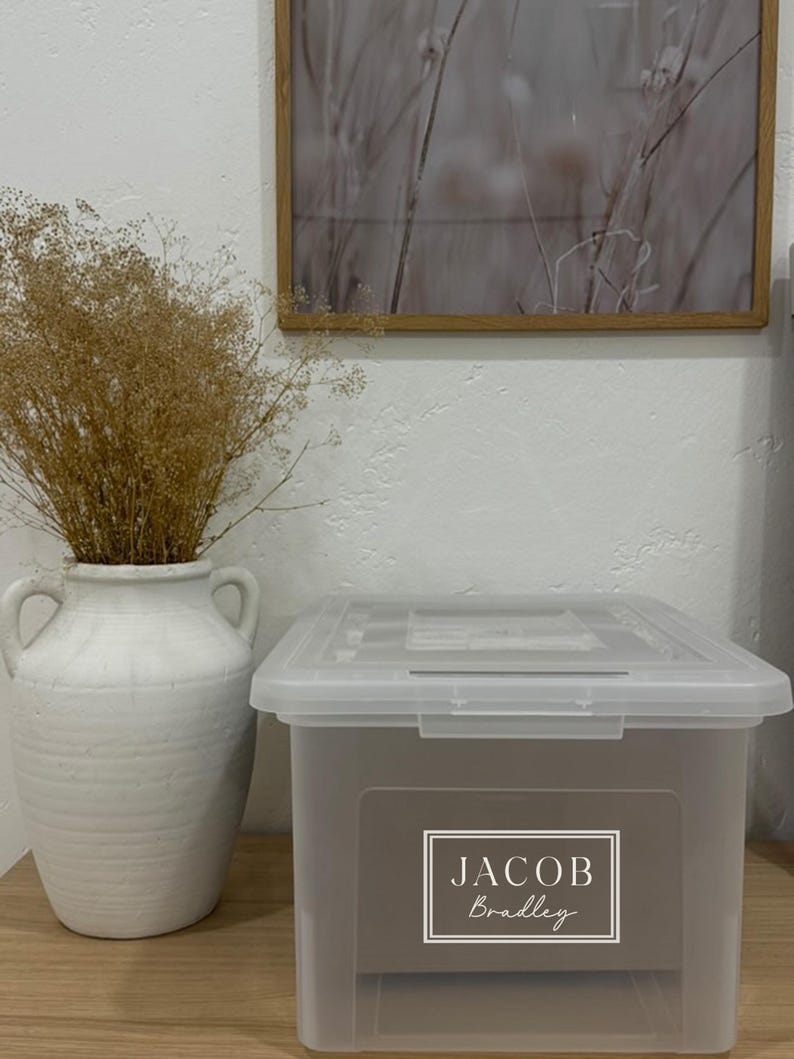 May include: A clear plastic storage bin with a gray front panel and lid. The front panel features the name "JACOB Bradley" in a white, elegant font within a white border. A white ceramic vase with handles holds dried brown plants.
