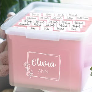 May include: A translucent pink plastic storage bin with a white lid, featuring a personalized white decal that reads Olivia ANN inside a geometric frame with a floral accent. The bin contains hanging file folders labeled with school grades.