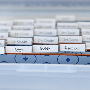 May include: A light blue storage container with labeled dividers. Labels include "Baby", "Toddler", "Preschool", and grade levels. The container has a blue and white patterned design.