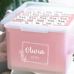 以下が含まれることがあります： ピンク色の収納ボックス。透明な蓋付きで、白い筆記体で「Olivia ANN」と書かれています。書類、スポーツ、記念品、幼稚園から12年生までの学年ラベルが付いたファイルフォルダーが入っています。整理整頓に最適です。