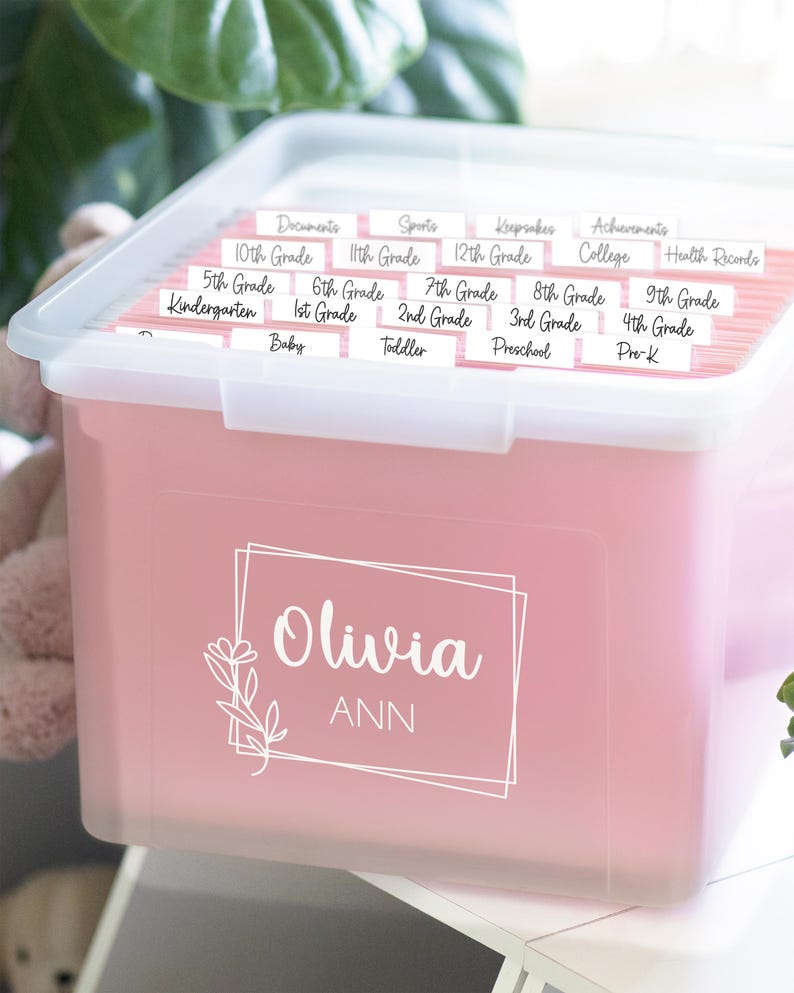 以下が含まれることがあります： ピンク色の収納ボックス。透明な蓋付きで、白い筆記体で「Olivia ANN」と書かれています。書類、スポーツ、記念品、幼稚園から12年生までの学年ラベルが付いたファイルフォルダーが入っています。整理整頓に最適です。