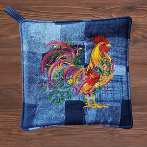 May include: A square potholder made of blue denim fabric with a patchwork pattern. The center features an intricate, colorful embroidery of a rooster with yellow, red, and purple feathers, surrounded by green vines and small floral accents.