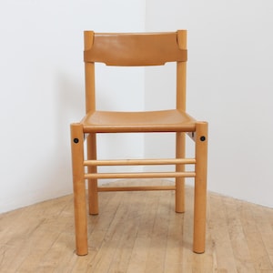 May include: A light-colored wooden chair featuring a tan leather seat and a matching tan leather backrest. The frame consists of four cylindrical legs connected by two horizontal support bars, with black circular hardware visible at the joints.
