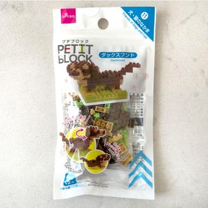 May include: A package of Petit Block building blocks, featuring a dachshund dog model. The package is white with pink and blue accents, and includes the text "PETIT BLOCK" and "Dachshund". The model is brown and tan, and sits on a green base.