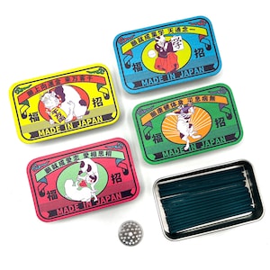 May include: Five rectangular, colourful Japanese incense boxes with illustrations and text. The boxes are in shades of yellow, blue, green, and red, with a small metal tray containing green incense sticks. A small, round metal incense holder is also present.