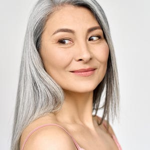 May include: A close-up portrait of a woman with long, straight, silver hair. She is looking to the side with a slight smile. She is wearing a pink spaghetti strap top. The background is a plain white.