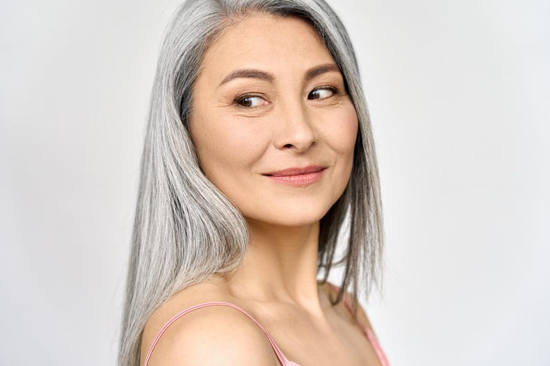 May include: A close-up portrait of a woman with long, straight, silver hair. She is looking to the side with a slight smile. She is wearing a pink spaghetti strap top. The background is a plain white.
