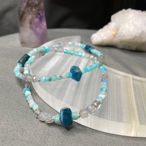Op de afbeelding: Twee armbanden met kralen van lichtblauwe, grijze en donkerblauwe edelstenen. De armbanden worden gepresenteerd op een wit, gebogen oppervlak. Een paars kristal en een wit kristal staan op de achtergrond.