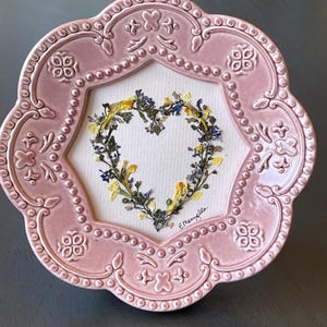 May include: A pink, ornate ceramic frame with a heart-shaped floral arrangement. The heart is made of yellow and blue dried flowers on a white background. The frame has a scalloped edge and decorative details.