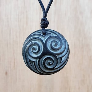 Könnte beinhalten: Eine schwarze Schnurkette mit einem runden Anhänger, der ein geschnitztes Design aus drei ineinandergreifenden Spiralen in hellblau und grün zeigt.