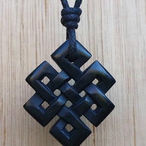 May include: Black wooden pendant necklace with a knotted design. The pendant is shaped like a square with rounded corners and has an intricate knot pattern. The necklace is made of black cord.