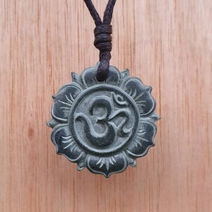 Puede incluir: Un collar con un colgante verde oscuro en forma de flor de loto con un símbolo Om tallado. El collar está hecho de un cordón marrón oscuro.