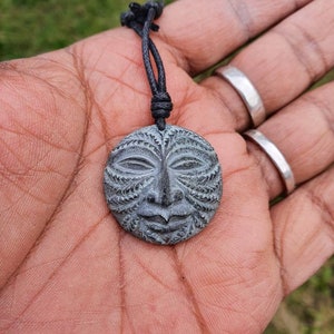 May include: A gray stone pendant with a carved face design, hanging from a black cord necklace.