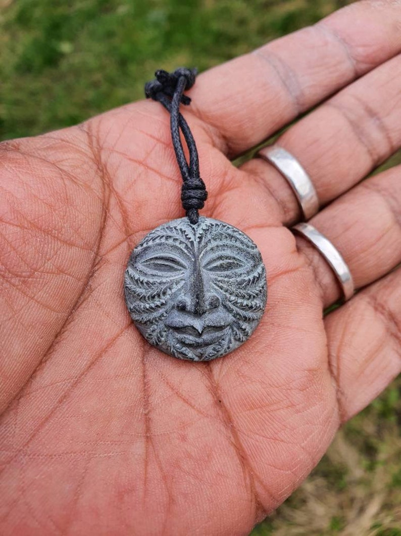 May include: A gray stone pendant with a carved face design, hanging from a black cord necklace.