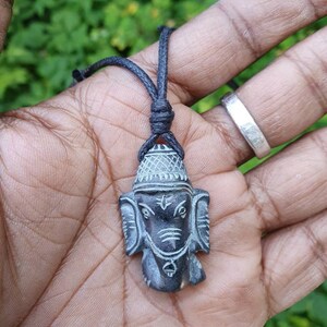 Pode incluir: Um pingente de pedra talhada em preto e cinza da divindade hindu Ganesha, em um cord&atilde;o preto.