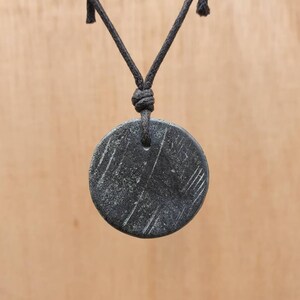 May include: A black round pendant with a textured surface hangs from a black cord necklace.