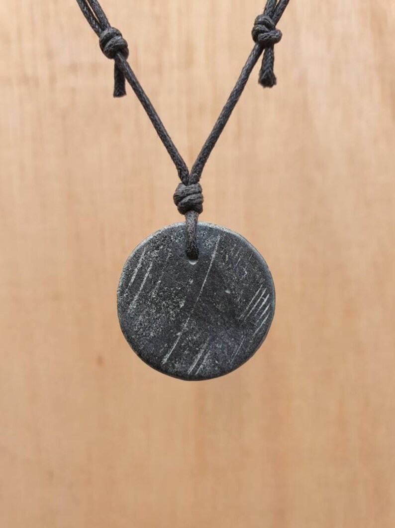 May include: A black round pendant with a textured surface hangs from a black cord necklace.