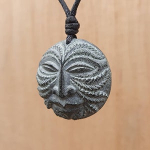 May include: A black cord necklace with a round, green and black stone pendant carved with a face. The face has a detailed design with leaves and a textured surface.
