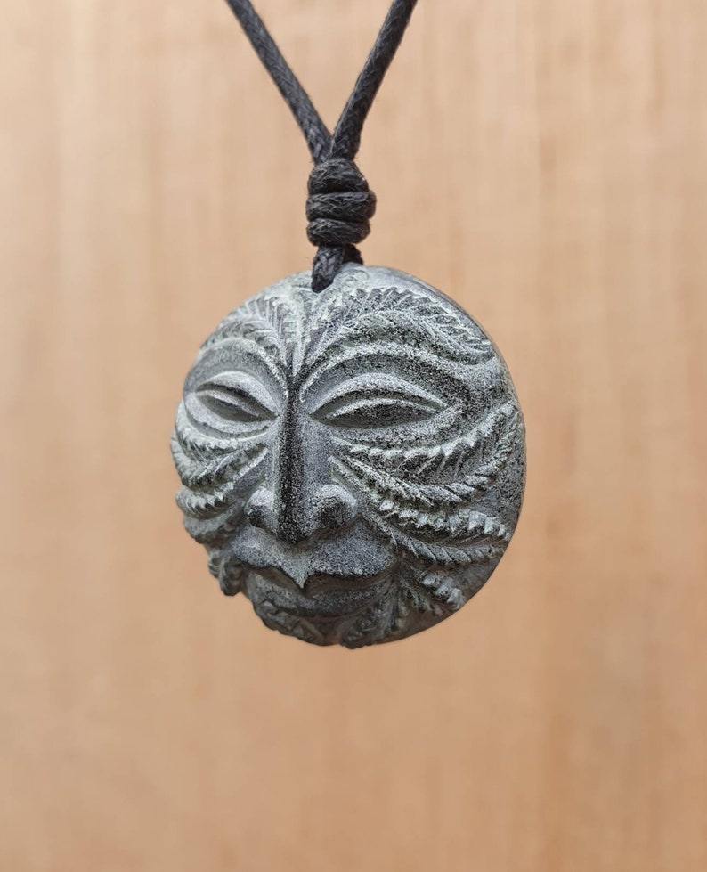 May include: A black cord necklace with a round, green and black stone pendant carved with a face. The face has a detailed design with leaves and a textured surface.
