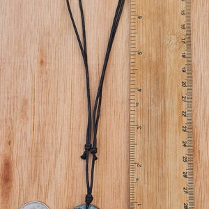 May include: A dark green stone pendant with a carved face design, hanging from a black cord. The pendant is about 1 inch in diameter and is shown next to a British one pound coin for size comparison.
