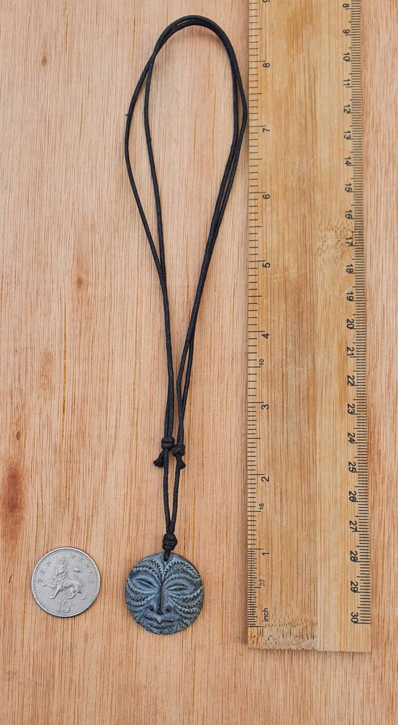 May include: A dark green stone pendant with a carved face design, hanging from a black cord. The pendant is about 1 inch in diameter and is shown next to a British one pound coin for size comparison.