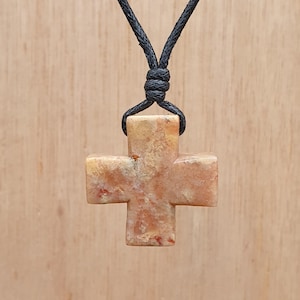 May include: A light brown stone cross pendant on a black cord necklace. The cross has a marbled pattern with hints of red and white.
