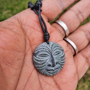 May include: A black cord necklace with a round, gray stone pendant featuring a carved face with intricate details.