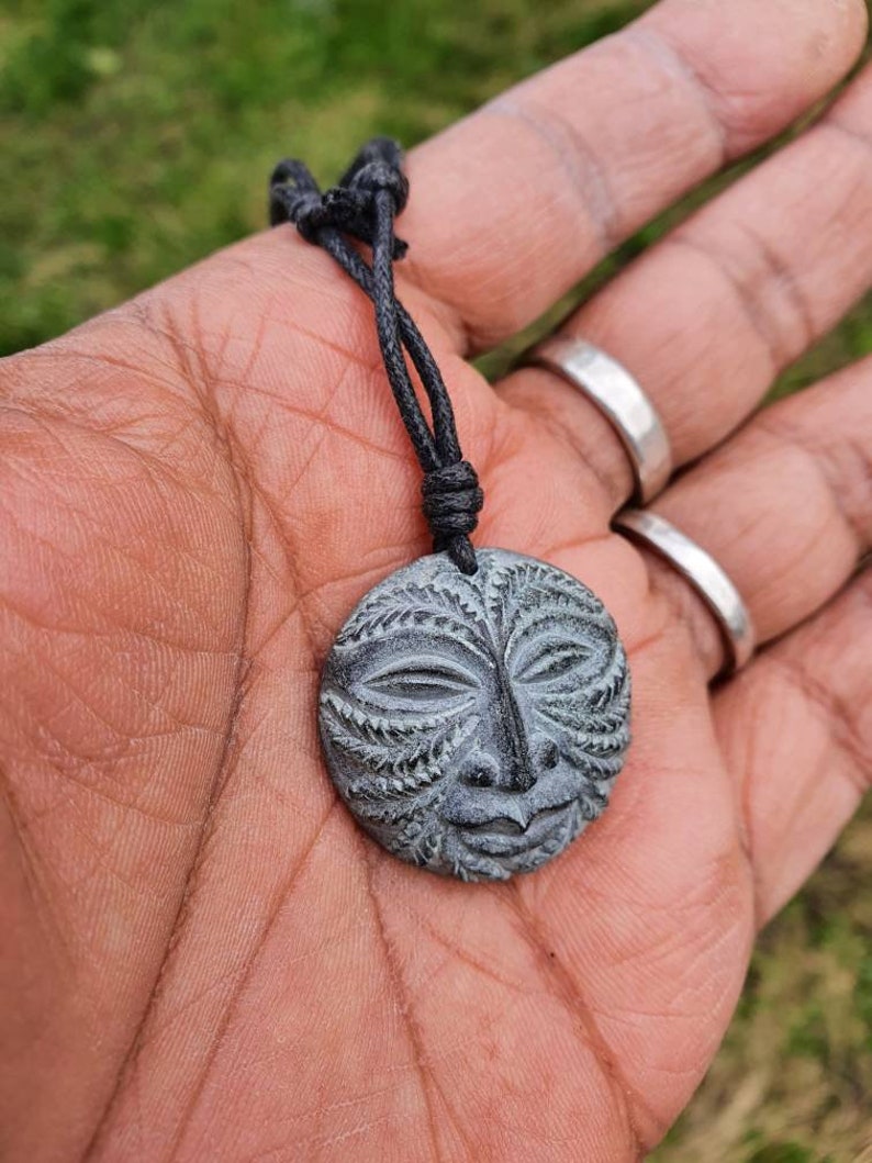 May include: A black cord necklace with a round, gray stone pendant featuring a carved face with intricate details.