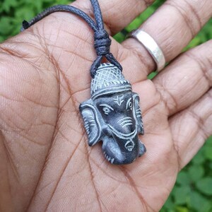 Pode incluir: Um pingente de pedra preta e cinza da divindade hindu Ganesha, esculpido em detalhe. O pingente est&aacute; preso a um cord&atilde;o preto.