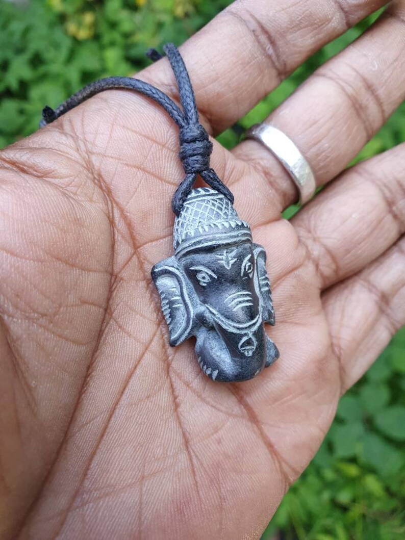 Pode incluir: Um pingente de pedra preta e cinza da divindade hindu Ganesha, esculpido em detalhe. O pingente est&aacute; preso a um cord&atilde;o preto.