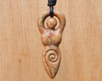 May include: A carved wooden pendant depicting a female figure with raised arms and a spiral design. The pendant is brown and beige with a natural wood grain. It is hanging from a black cord.
