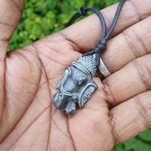 Pode incluir: Um pingente de pedra talhada em preto e cinza da divindade hindu Ganesha, pendurado em um cord&atilde;o preto.