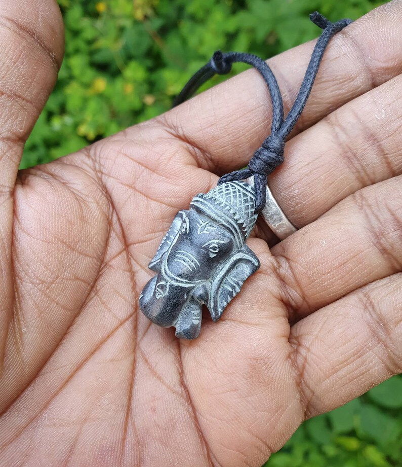 Pode incluir: Um pingente de pedra talhada em preto e cinza da divindade hindu Ganesha, pendurado em um cord&atilde;o preto.