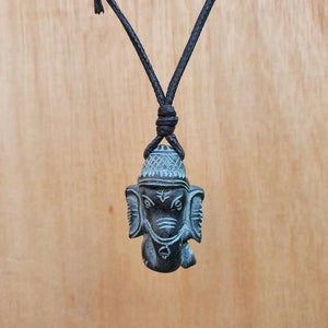 Pode incluir: Um colar de cord&atilde;o preto com um pingente de pedra cinza escuro esculpido com a imagem de Ganesha, o deus hindu dos come&ccedil;os, do sucesso e da boa sorte.