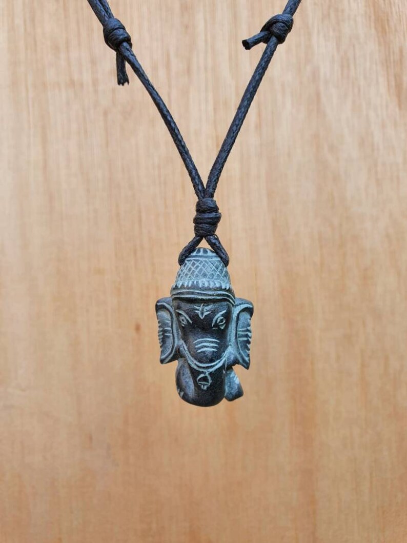 Pode incluir: Um colar de cord&atilde;o preto com um pingente de pedra cinza escuro esculpido com a imagem de Ganesha, o deus hindu dos come&ccedil;os, do sucesso e da boa sorte.