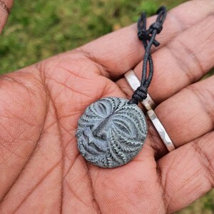 May include: A dark gray carved wooden pendant with a face design, hanging from a black cord.
