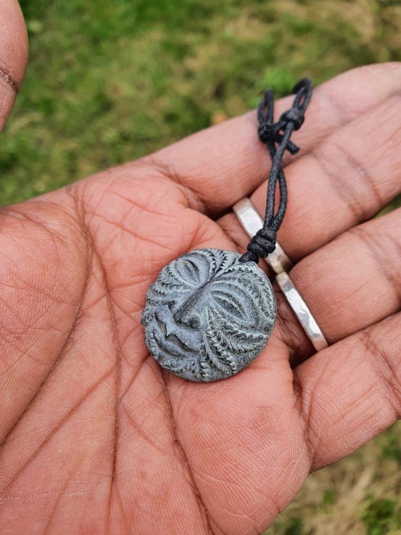 May include: A dark gray carved wooden pendant with a face design, hanging from a black cord.