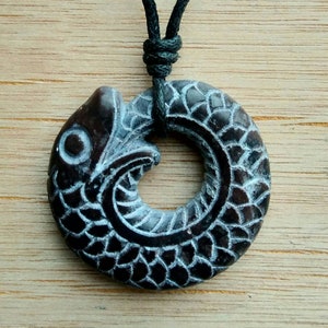 May include: A black wooden pendant necklace featuring a carved fish design. The fish is depicted in a circle shape with its tail wrapping around its head. The pendant is attached to a black cord.
